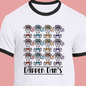 May include: White t-shirt with a black ringer neck featuring a pattern of 12 cartoon characters wearing hats and holding musical notes. The text "The Dapper Dan's" is printed below the characters.