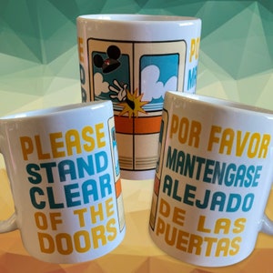 May include: Three white ceramic mugs with a Disney theme. The mugs feature a cartoon image of Mickey Mouse's hand reaching out of a train window. The text on the mugs reads "Please Stand Clear of the Doors" and "Por Favor Mantenga Se Alejado De Las Puertas".