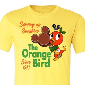 May include: Yellow t-shirt with a cartoon orange bird holding a Mickey Mouse ice cream bar. The text on the shirt reads "Serving up Sunshine The Orange Bird Since 1971".