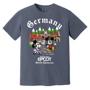May include: A blue t-shirt with a white graphic of Mickey and Minnie Mouse in German attire in front of a German village. The text "Germany" is above the graphic and "Walt Disney World EPCOT World Showcase" is below the graphic.