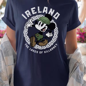 Puede incluir: Camiseta azul marino con Mickey Mouse de dibujos animados con gorro y pantalones cortos a cuadros verdes, con el texto "IRELAND" y "THE LAKES OF KILLARNEY" en un dise&ntilde;o circular blanco. La camiseta tiene mangas enrolladas.
