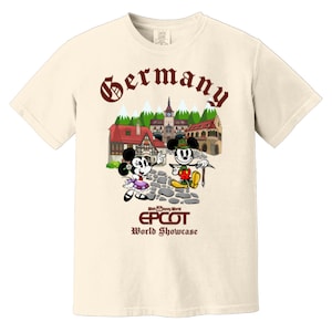 May include: A beige t-shirt with a brown graphic of Mickey and Minnie Mouse in German attire. The graphic includes a small village scene with the text "Germany" above it and "Walt Disney World EPCOT World Showcase" below it.