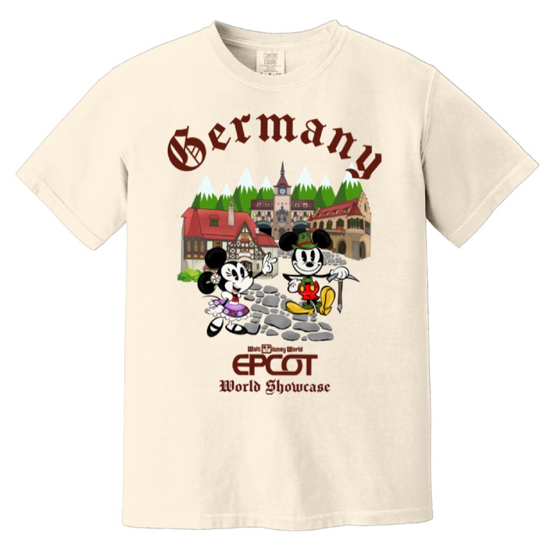 May include: A beige t-shirt with a brown graphic of Mickey and Minnie Mouse in German attire. The graphic includes a small village scene with the text "Germany" above it and "Walt Disney World EPCOT World Showcase" below it.
