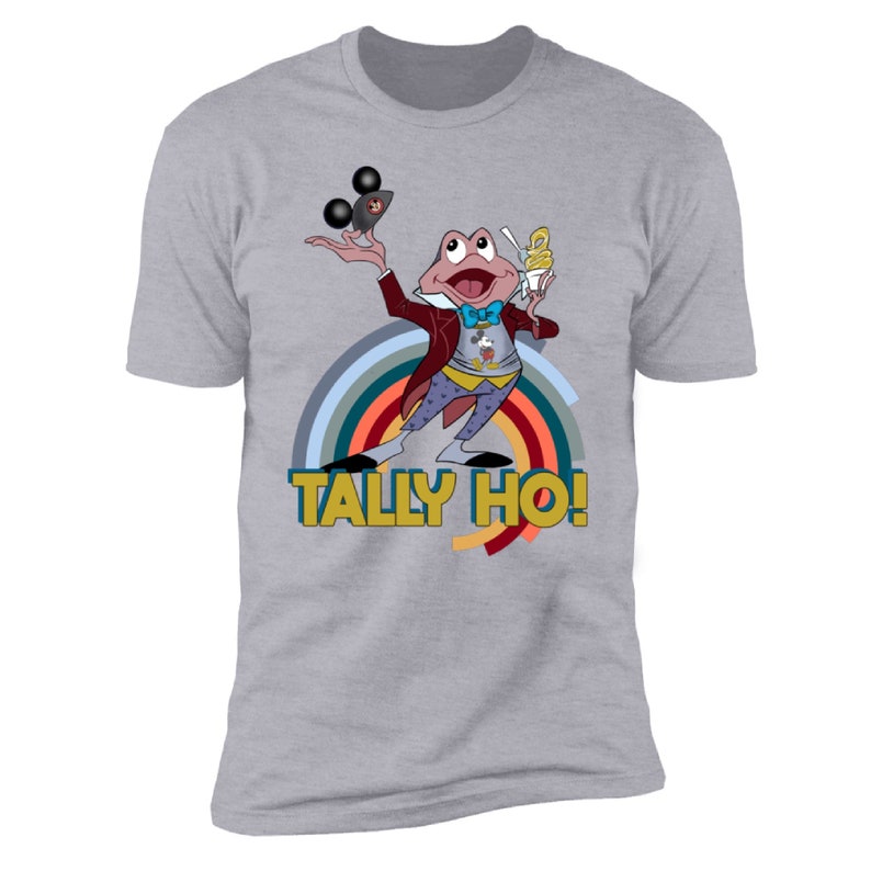 May include: A light grey t-shirt with a cartoon frog character holding a Mickey Mouse-shaped object and an ice cream cone. The frog is wearing a burgundy jacket, a blue bow tie, and a vest with a Mickey Mouse design. The text "TALLY HO!" is below the frog.