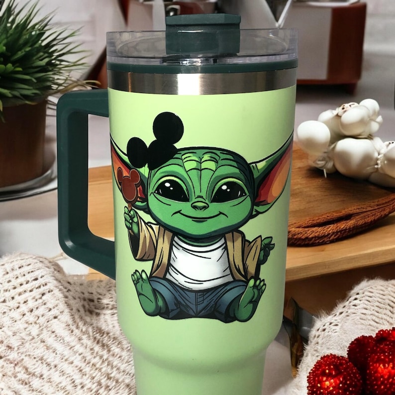 Op de afbeelding: Een groene reistumbler van roestvrij staal met een groen deksel en handvat. De tumbler heeft een cartoonafbeelding van een groen buitenaards wezen dat een Mickey Mouse-oor-ijsje vasthoudt. Het buitenaardse wezen draagt een bruin jack, een wit shirt en een blauwe spijkerbroek.