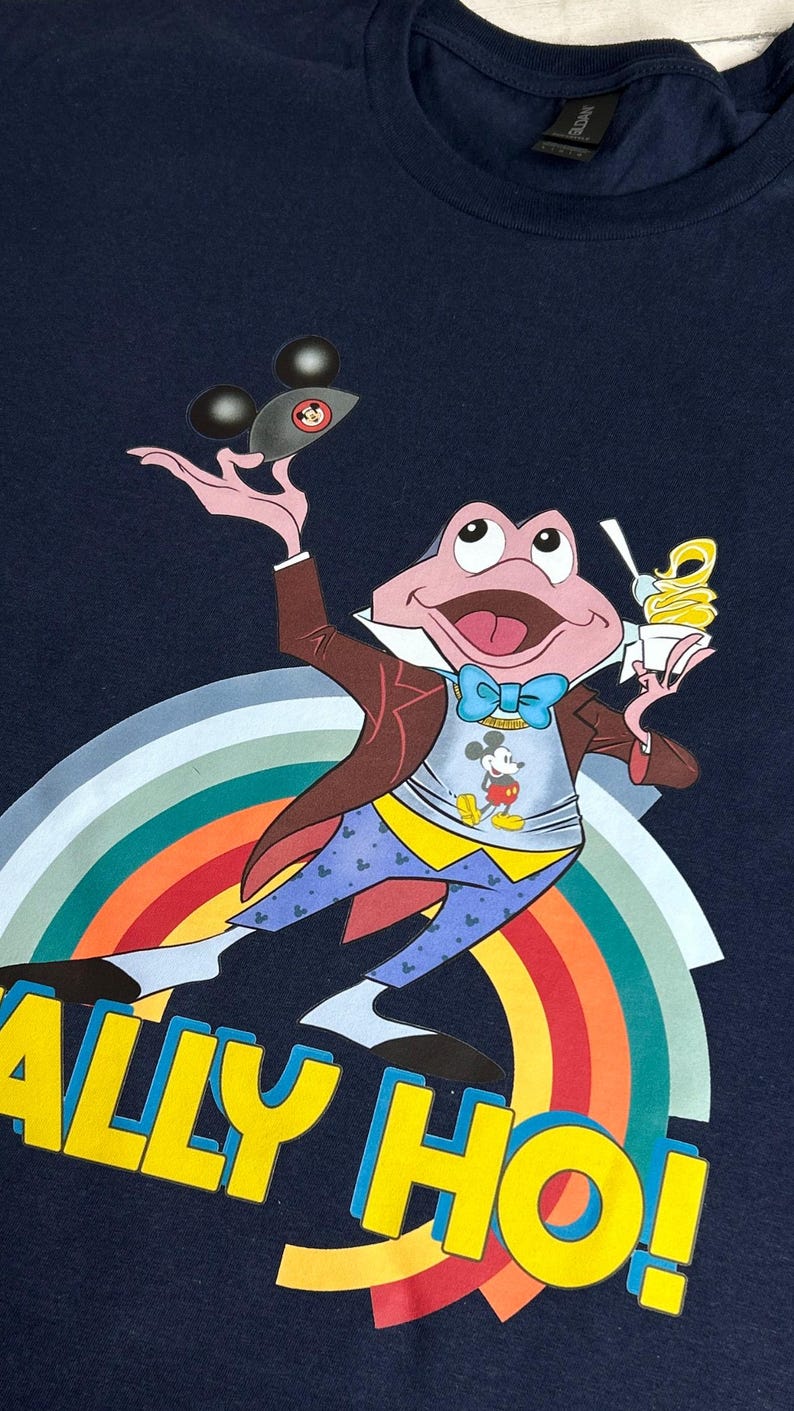 May include: Navy blue t-shirt with a cartoon frog character holding a Mickey Mouse-shaped object and ice cream. The frog wears a suit and bow tie, with a Mickey Mouse shirt. The shirt has the text "ALLY HO!" in yellow, above a rainbow.