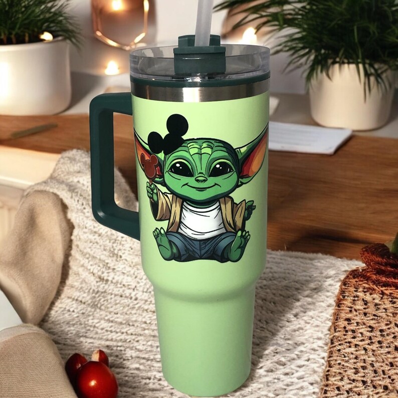 Op de afbeelding: Groene reistumbler van roestvrij staal met een groene deksel en een handvat. De tumbler heeft een cartoonafbeelding van een groen buitenaards wezen dat een bruin jack en een blauwe spijkerbroek draagt. Het buitenaardse wezen draagt een Mickey Mouse-oorkap.