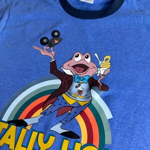 May include: Blue t-shirt with a navy blue collar. It features a cartoon frog character holding ice cream and a Mickey Mouse-themed object. The frog is wearing a suit and bow tie, with a rainbow and the words "TALLY HO!" in yellow.