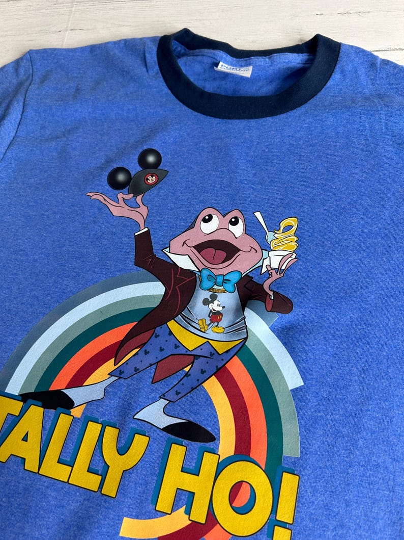 May include: Blue t-shirt with a navy blue collar. It features a cartoon frog character holding ice cream and a Mickey Mouse-themed object. The frog is wearing a suit and bow tie, with a rainbow and the words "TALLY HO!" in yellow.