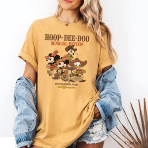 Könnte beinhalten: Senffarbenes T-Shirt mit einer Grafik von Mickey Mouse, Donald Duck und anderen Charakteren in Cowboy-Kleidung. Das Shirt hat den Text "HOOP-DEE-DOO MUSICAL REVIEW" und "FORT WILDERNESS RESORT Walt Disney World" aufgedruckt.