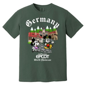 May include: A green t-shirt with a white graphic of Mickey and Minnie Mouse in German attire in front of a German village. The text "Germany" is above the image and "Walt Disney World EPCOT World Showcase" is below the image.