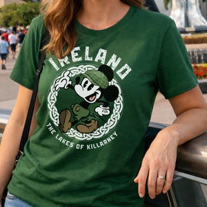 Puede incluir: Camiseta verde con Mickey Mouse vestido con atuendo irland&eacute;s, con la palabra "IRELAND" sobre el personaje. El dise&ntilde;o incluye un borde de nudo celta y el texto "THE LAKES OF KILLARNEY".