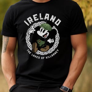 Puede incluir: Camiseta negra con un personaje de dibujos animados con un sombrero verde y un objeto verde, dentro de un dise&ntilde;o de nudo celta blanco. Los textos "IRELAND" y "THE LAKES OF KILLARNEY" est&aacute;n presentes.