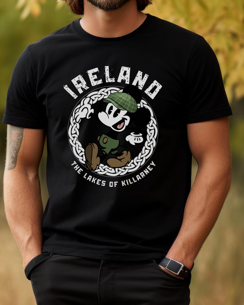 Puede incluir: Camiseta negra con un personaje de dibujos animados con un sombrero verde y un objeto verde, dentro de un dise&ntilde;o de nudo celta blanco. Los textos "IRELAND" y "THE LAKES OF KILLARNEY" est&aacute;n presentes.
