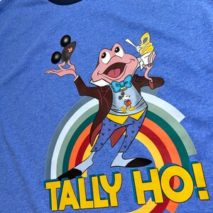 May include: Blue ringer t-shirt with a cartoon frog character wearing a bow tie, holding a black car and an ice cream cone. The frog is in front of a rainbow, with the words "TALLY HO!" in yellow.