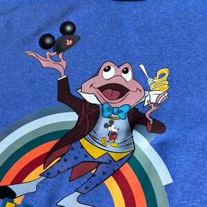 May include: Blue ringer t-shirt with a cartoon frog character holding a Mickey Mouse ice cream and a Mickey Mouse hat. The frog is wearing a suit and bow tie, with a rainbow and the words "LLY HO!" in yellow.