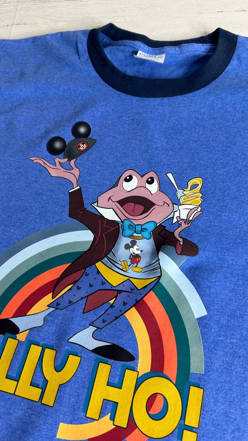 May include: Blue ringer t-shirt with a cartoon frog character holding a Mickey Mouse ice cream and a Mickey Mouse hat. The frog is wearing a suit and bow tie, with a rainbow and the words "LLY HO!" in yellow.