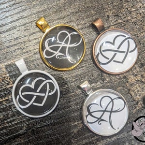 May include: Four round pendants with different colored metal frames. Each pendant features a black and white graphic of an infinity symbol intertwined with a heart. The pendants are on a wooden surface.