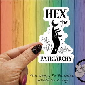 May include: A white sticker with black text and a black silhouette of a hand with a crescent moon and stars. The text reads "HEX the PATRIARCHY".