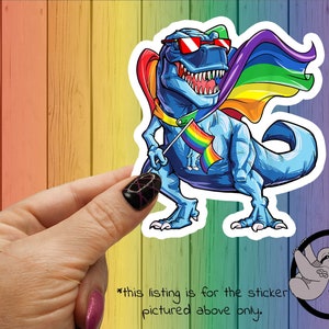 May include: A blue dinosaur wearing sunglasses and a rainbow cape holds a rainbow pride flag. The dinosaur is standing on a wooden background.