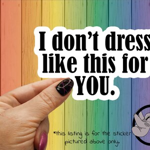 May include: A white sticker with black text that reads "I don't dress like this for YOU." The sticker is being held by a hand with black and pink nail polish. The background is a rainbow colored wood panel.
