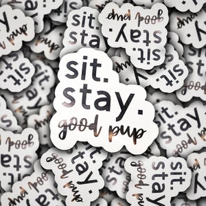 May include: A close-up of several white stickers with black text that reads "sit. stay. good pup". The stickers are arranged in a random pattern and have a slightly distressed, vintage look.