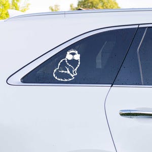 May include: A white vinyl decal applied to a car window, featuring a stylized illustration of a fluffy cat wearing a small crown and large sunglasses. The cat is depicted in a sitting position with a long, bushy tail and distinct fur outlines.