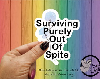 May include: A sticker with a watercolor design of blue flowers and the text "Surviving Purely Out Of Spite". The sticker is held by a hand with black and pink nail polish. The sticker is on a rainbow colored wooden background.
