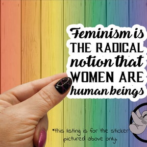 May include: A sticker with the text "Feminism is THE RADICAL notion that WOMEN ARE human beings". The sticker is on a rainbow colored wooden background.
