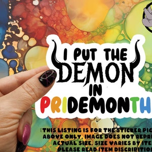 May include: A sticker with the text "I put the demon in pride month" in black lettering with rainbow colored letters for "pride month". The sticker is on a colorful background.