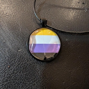 May include: A round pendant necklace featuring the non-binary pride flag design. The pendant has a black border and hangs from a black cord. The flag design includes yellow, white, purple, and black stripes.