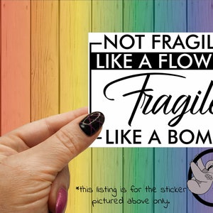 May include: A white sticker with black text that reads "Not Fragile Like A Flower Fragile Like A Bomb". The sticker is being held by a hand with pink and black nail polish.
