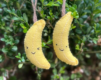 May include: Two yellow felt banana ornaments with stitched details and embroidered faces. Each banana features a simple smiling expression and is suspended by a thin brown twine loop, designed for hanging display with visible white thread stitching.