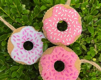Puede incluir: Tres adornos de fieltro con forma de donut con glaseado y chispas rosas y blancas, cada uno con una base beige y una cuerda de yute para colgar. Los donuts están colocados sobre un fondo de hojas verdes.