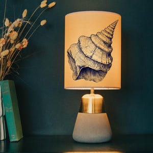 May include: A table lamp with a beige lampshade, decorated with a seashell design. It's on a dark blue dresser, beside a vase of dried flowers and a green book. The room has a dark teal wall. The lamp is a decorative item.