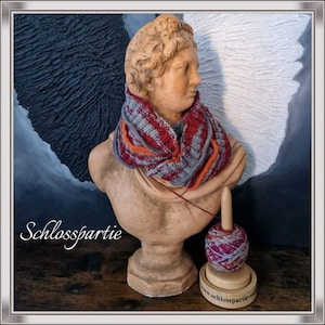 Puede incluir: Un busto de una persona que lleva una bufanda infinita a rayas grises, rojas y naranjas. El busto está sobre una mesa de madera con un devanador de hilo de madera y una madeja de hilo de color rojo, gris y naranja. El devanador de hilo tiene una etiqueta que dice "www.schlosspartie.web".