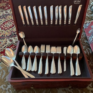May include: A wooden silverware case with a dark red finish, open to reveal a set of silver utensils. The set includes knives, forks, and spoons, neatly arranged in a dark brown velvet-lined interior. The brand name "Pacific Silvercloth" is visible.