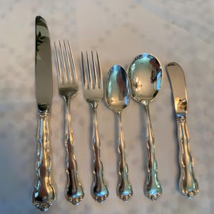 May include: A six-piece silver-toned cutlery set comprising a dinner knife, two forks of different sizes, two spoons of different sizes, and a butter knife. Each item has a polished finish and a decorative, scalloped handle design.