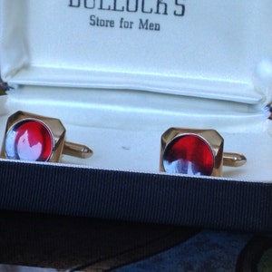 May include: A pair of gold-toned cufflinks with a square design and a vibrant red center. The cufflinks are presented in a white box with the text "BULLOCK'S Store for Men" visible on the lid.