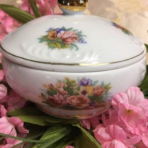 May include: A white porcelain trinket box with a gold lid and floral design. The box is decorated with pink, yellow, and blue flowers.