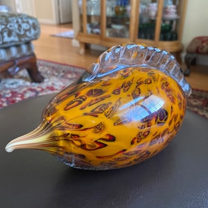 May include: A glass sculpture of a bird with a yellow, orange, and brown marbled design. The bird has a long, thin beak and a rounded body.