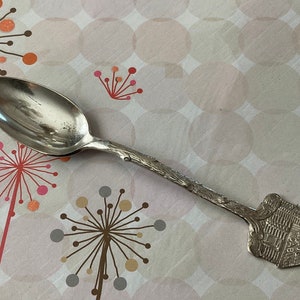 May include: A silver spoon with a detailed design of a building on the handle. The spoon is lying on a pink and white patterned surface.