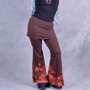 May include: A brown, tie-dye, wide-leg trouser with a brown, fitted, short skirt overlay. The trouser has a red and brown tie-dye pattern on the bottom of the legs. The model is wearing a black tank top with a lace-up detail in the front.
