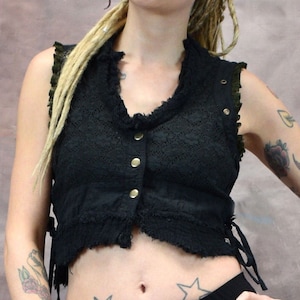 Könnte beinhalten: Ein schwarzes Spitzen-Crop-Top mit Knopfverschluss und verstellbaren Trägern. Das Top wird von einer Person mit blonden Dreadlocks und Tattoos getragen. Die Person trägt eine schwarze Hose und hat ein Bauchnabelpiercing.