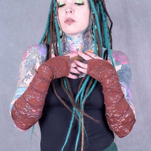 May include: A person with teal and brown dreadlocks is wearing a black tank top and a pair of brown lace arm warmers. They have several tattoos on their arms and neck.