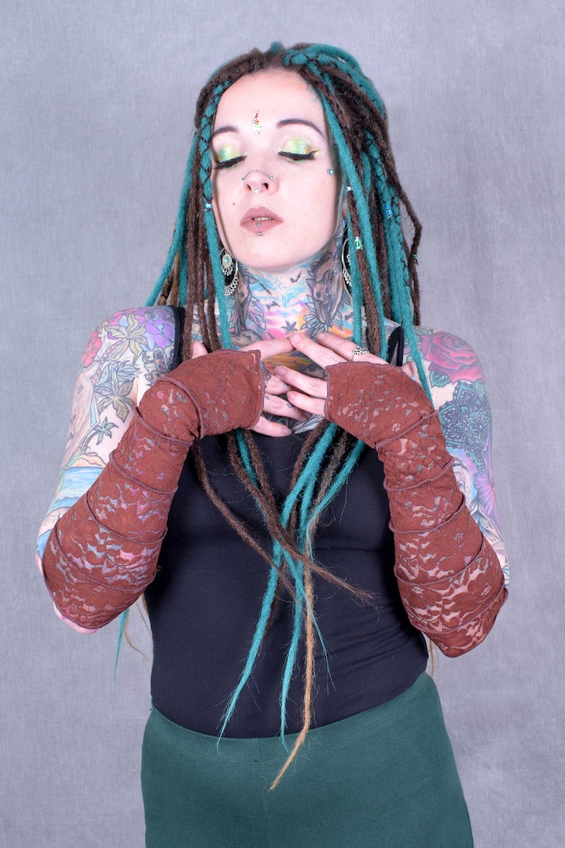 May include: A person with teal and brown dreadlocks is wearing a black tank top and a pair of brown lace arm warmers. They have several tattoos on their arms and neck.