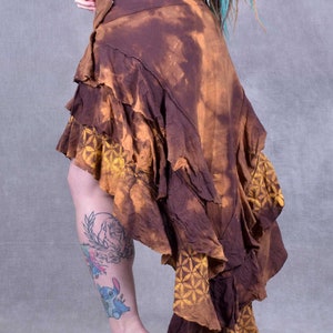 May include: A brown and yellow asymmetrical skirt with a ruffled hem and a tie closure. The skirt is made of a soft, flowing fabric and has a tie closure at the waist.