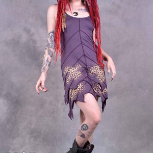 Może przedstawiać: Fioletowa, bezrękawowa sukienka z geometrycznym wzorem i strzępionymi krawędziami. Sukienkę nosi osoba z czerwonymi włosami i tatuażami. Osoba nosi czarne buty.