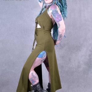 A woman wearing a green maxi dress with a lace-up front and side slits. She has blue and green dreadlocks and tattoos on her arms and legs. She is wearing black boots with buckles.