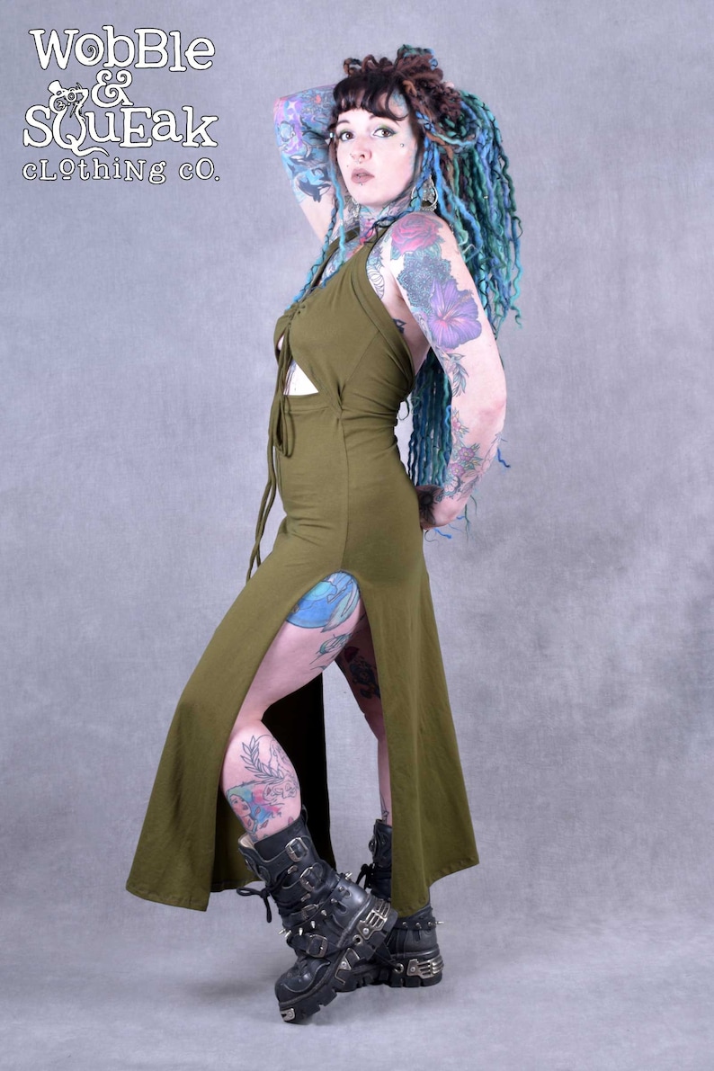 A woman wearing a green maxi dress with a lace-up front and side slits. She has blue and green dreadlocks and tattoos on her arms and legs. She is wearing black boots with buckles.